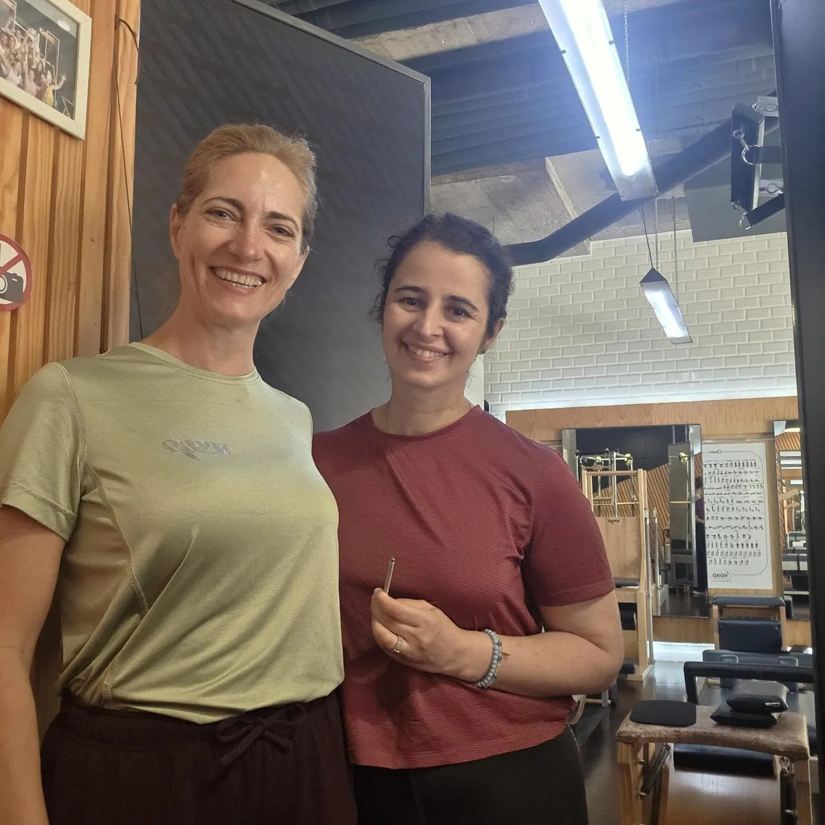 Duas instrutoras do estúdio, sorridentes, com a sala de aparelhos atrás