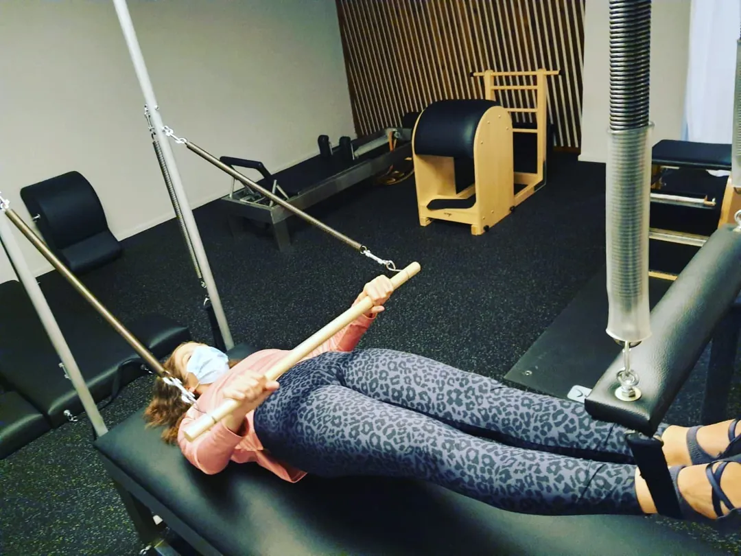 Aluna deitada no reformer a puxar as correias, com uma chair ao fundo
